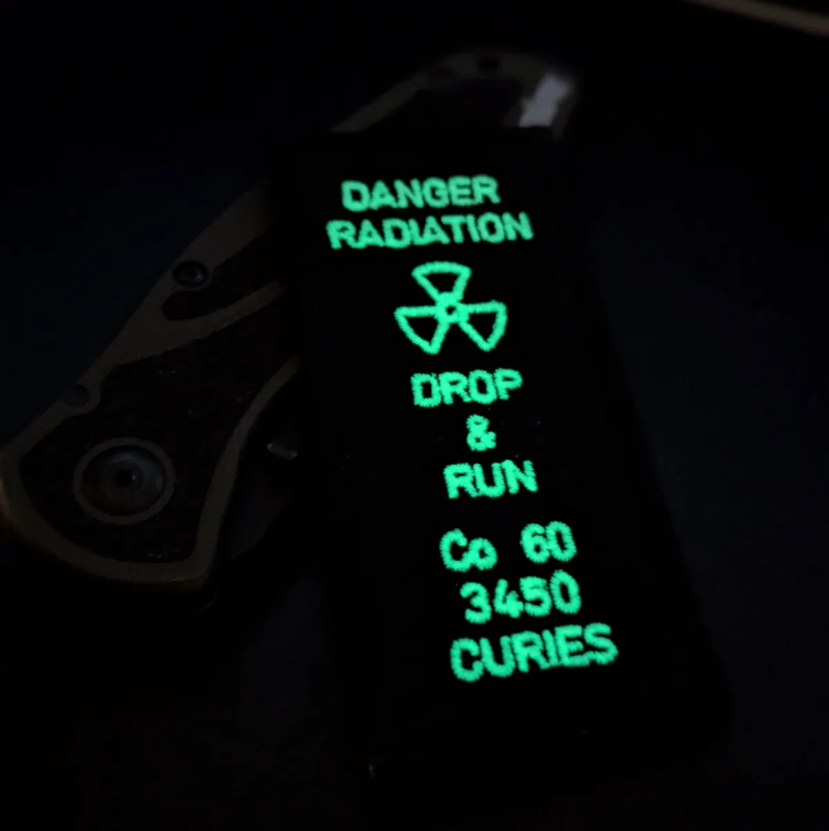 A dark image shows a glowing green patch with radiation warning text and a trefoil symbol, placed next to a folding knife…