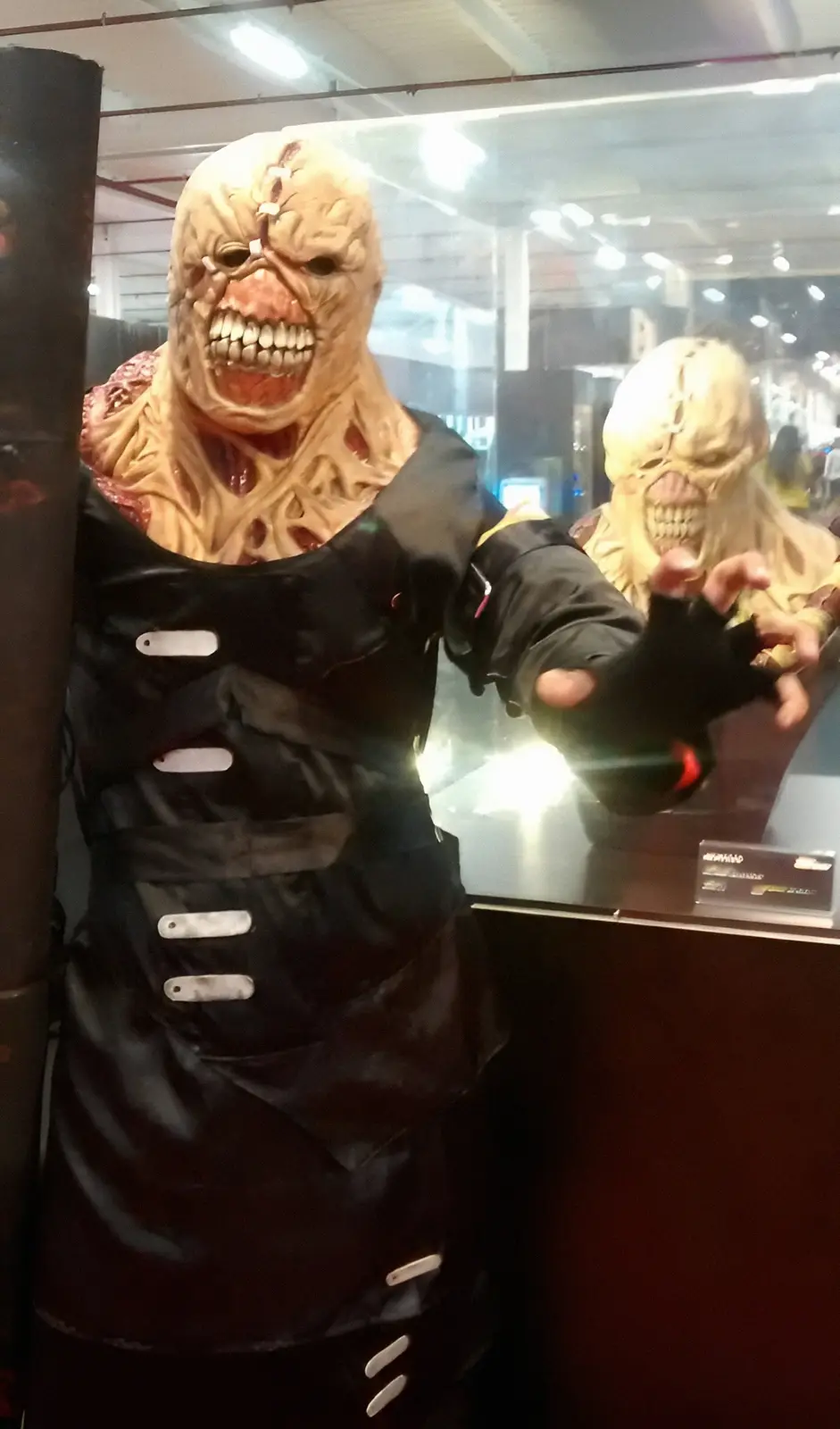 A person in a grotesque, flesh-torn monster costume with a black apron stands behind a glass display case, reaching out with…