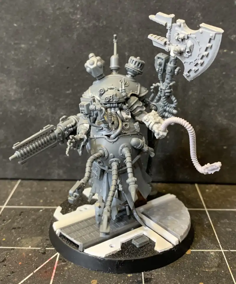 A grey, unpainted miniature of a heavily armed, multi-limbed sci-fi warrior stands on a base, with overlaid text reading…