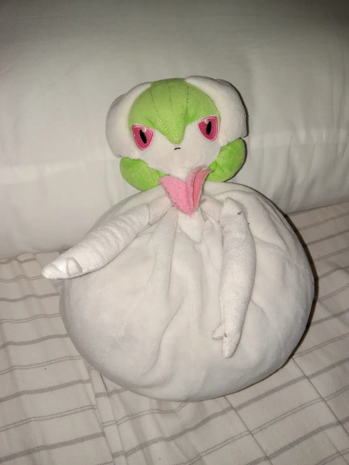 A plush toy of the Pokémon Goomy, featuring a green head, pink eyes, and a white body, is shown sitting on a striped…