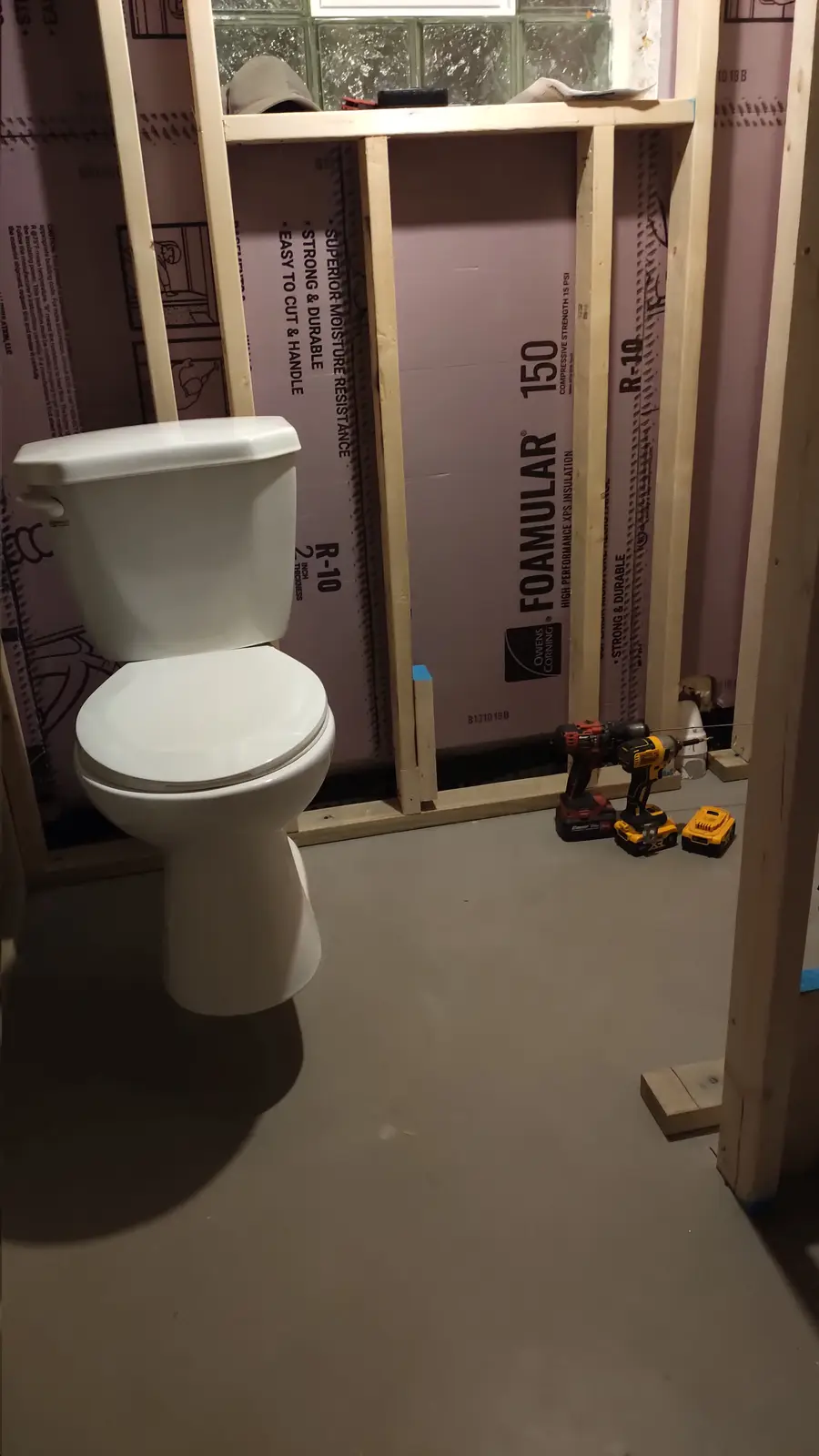 A white toilet sits in an unfinished bathroom with exposed wooden framing and pink Owens Corning Foamular 150 insulation…