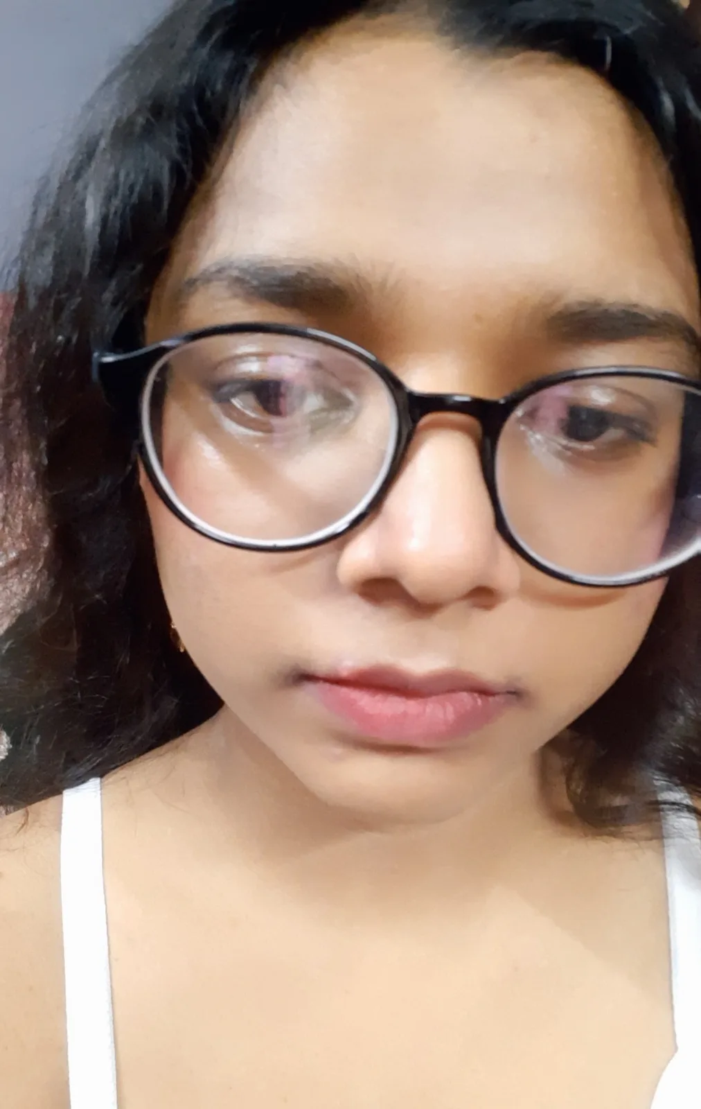 A close-up selfie of a person with dark curly hair and black-rimmed glasses, wearing a white tank top, looking directly at…