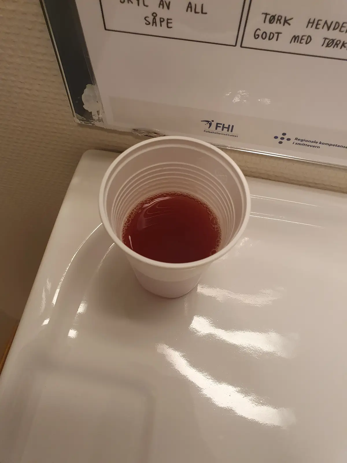 A white plastic cup filled with reddish-brown liquid sits in a white sink, with a sign above in Norwegian reading 'SKYL AV…