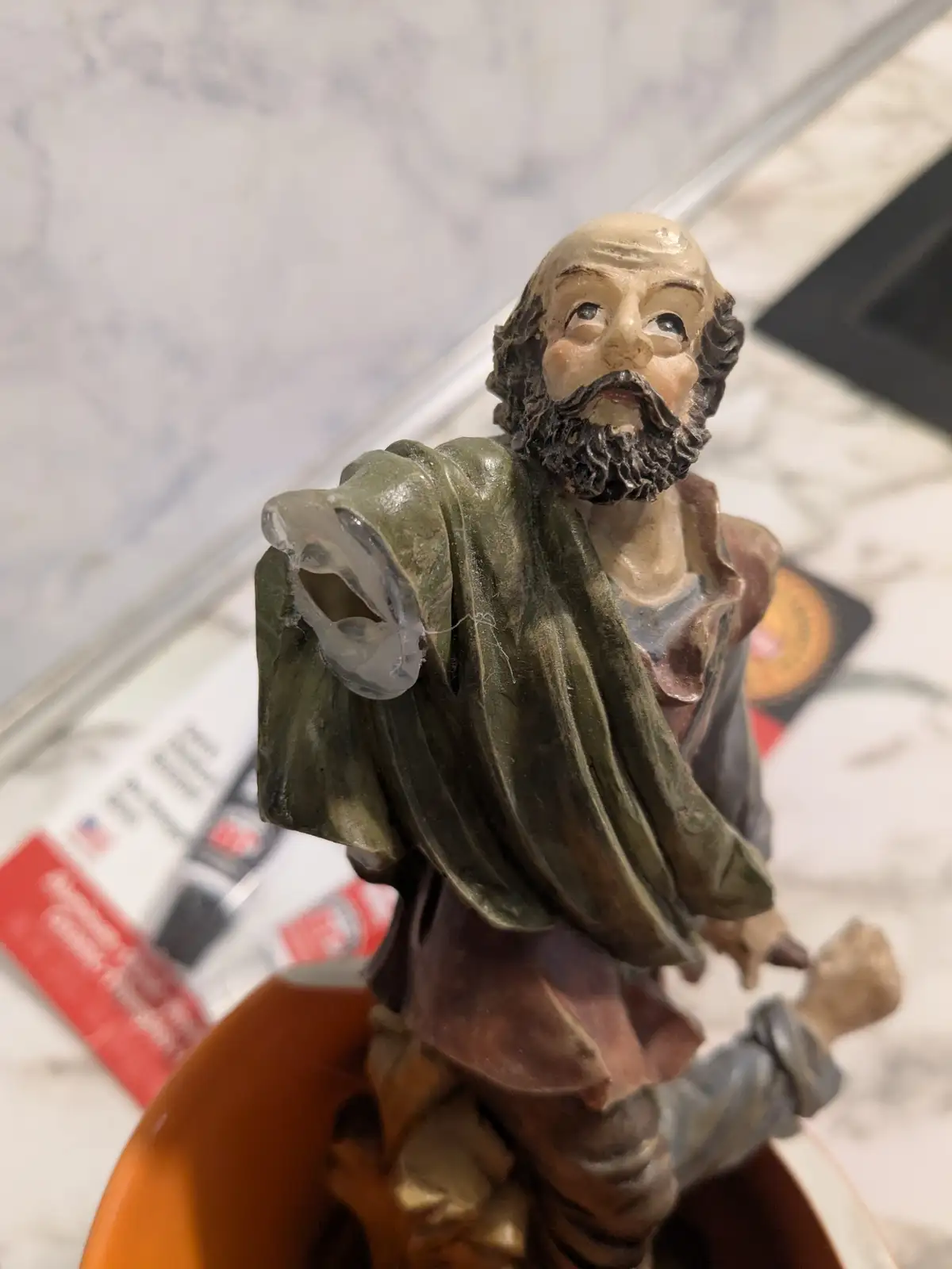 A close-up photo shows a small, painted figurine of a bearded man in robes, likely a religious figure, standing on an orange…