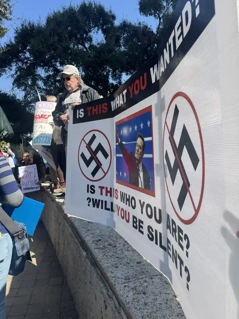 A protest sign features a photo of Elon Musk in a campaign-style frame, flanked by two swastikas crossed out in red circles…