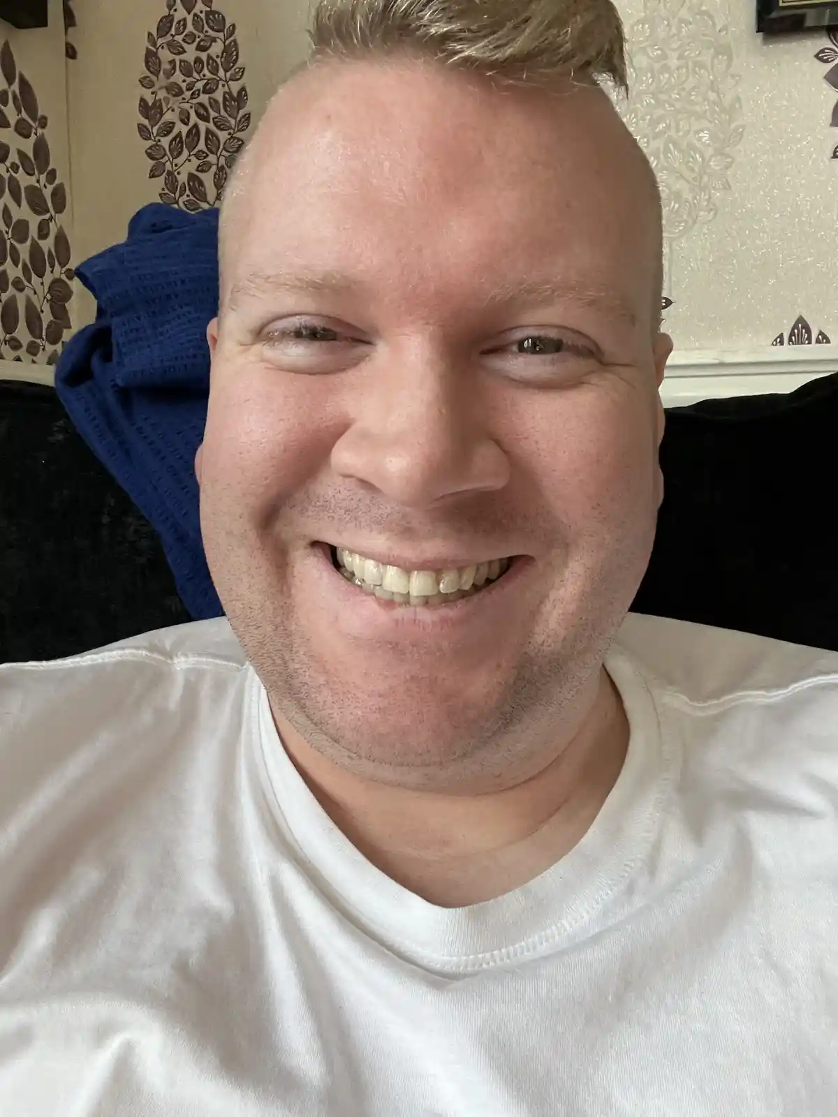 A close-up selfie of a smiling man with short blond hair and a white t-shirt, sitting on a dark couch with a blue blanket…