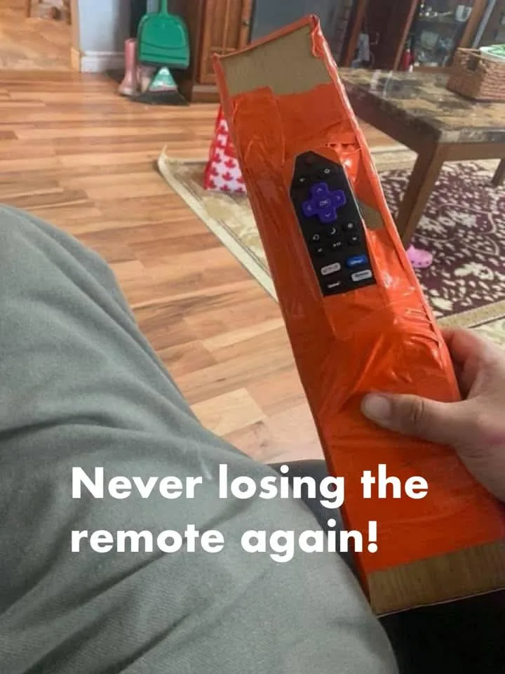 A person holds a television remote control that is taped to a long, orange cardboard tube, making it bulky and hard to lose…