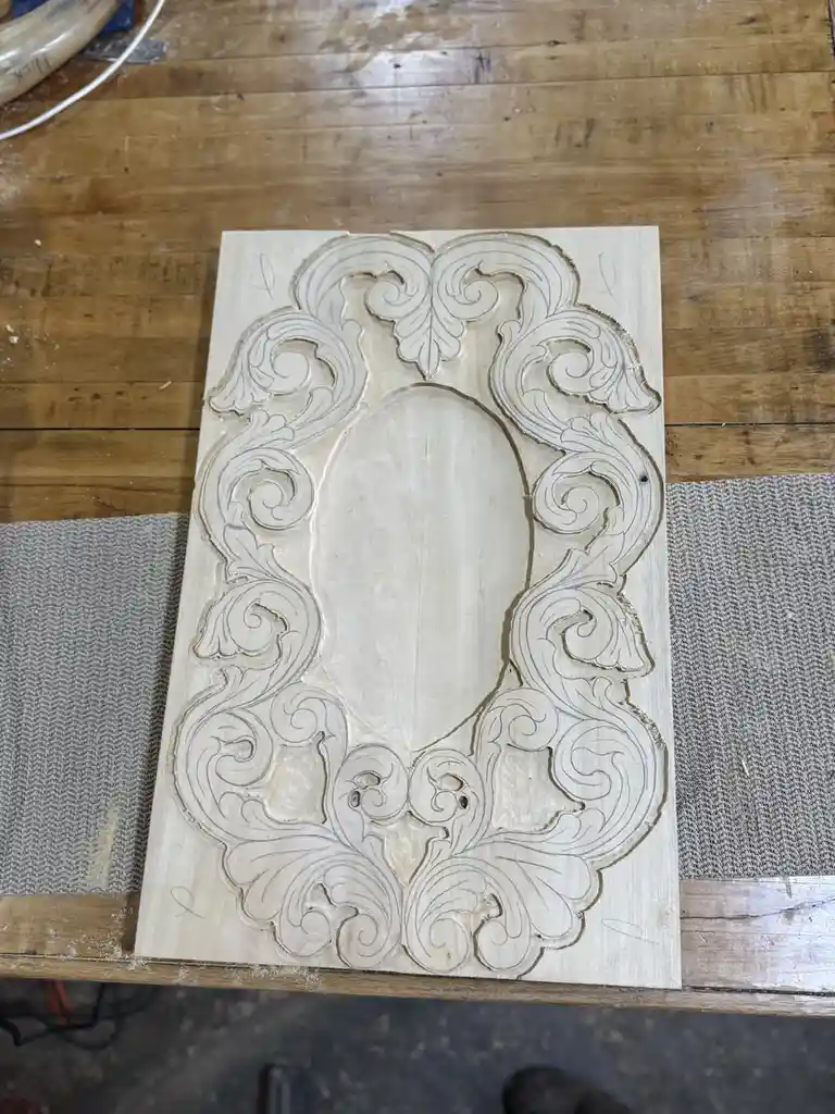 A piece of light-colored wood with intricate, symmetrical scrollwork carved into it, resembling a decorative frame or mirror…