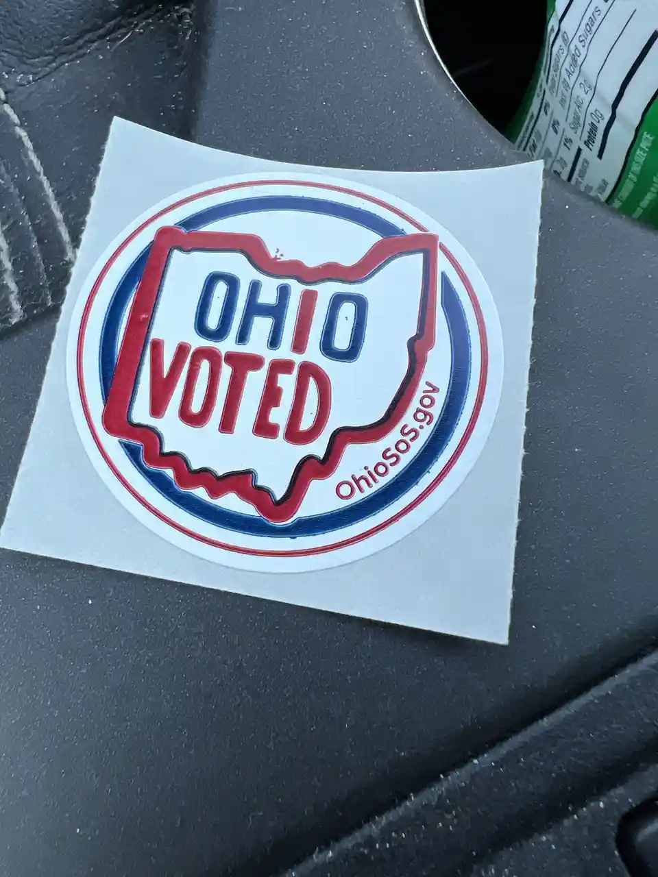 A close-up photo shows an official-looking circular sticker with the outline of the state of Ohio in red, containing the…
