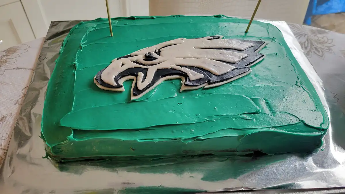 A rectangular cake with green frosting and a white and black Philadelphia Eagles logo on top, sitting on aluminum foil. Two…