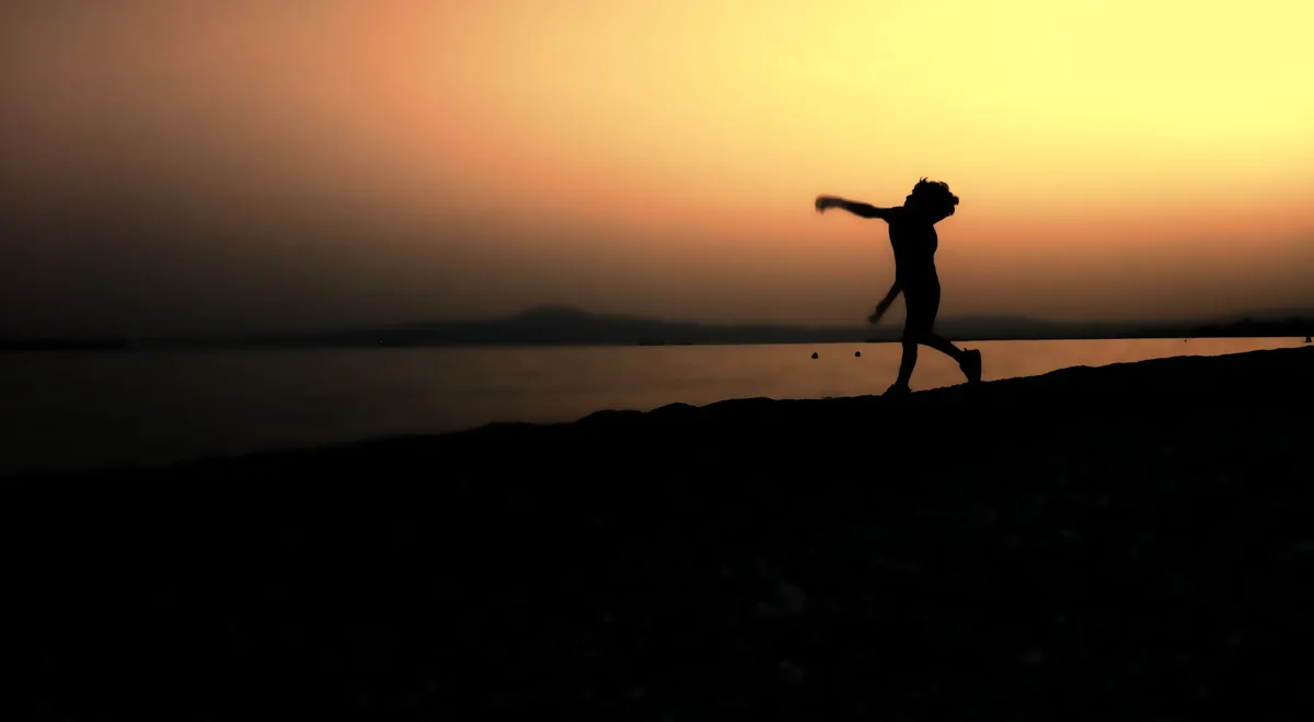 A silhouette of a person joyfully jumping on a beach at sunset, arms outstretched against a warm orange and yellow sky. The…