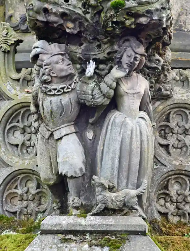 A weathered stone sculpture depicts a man and woman in historical attire, both looking upward with hands raised, as a small…