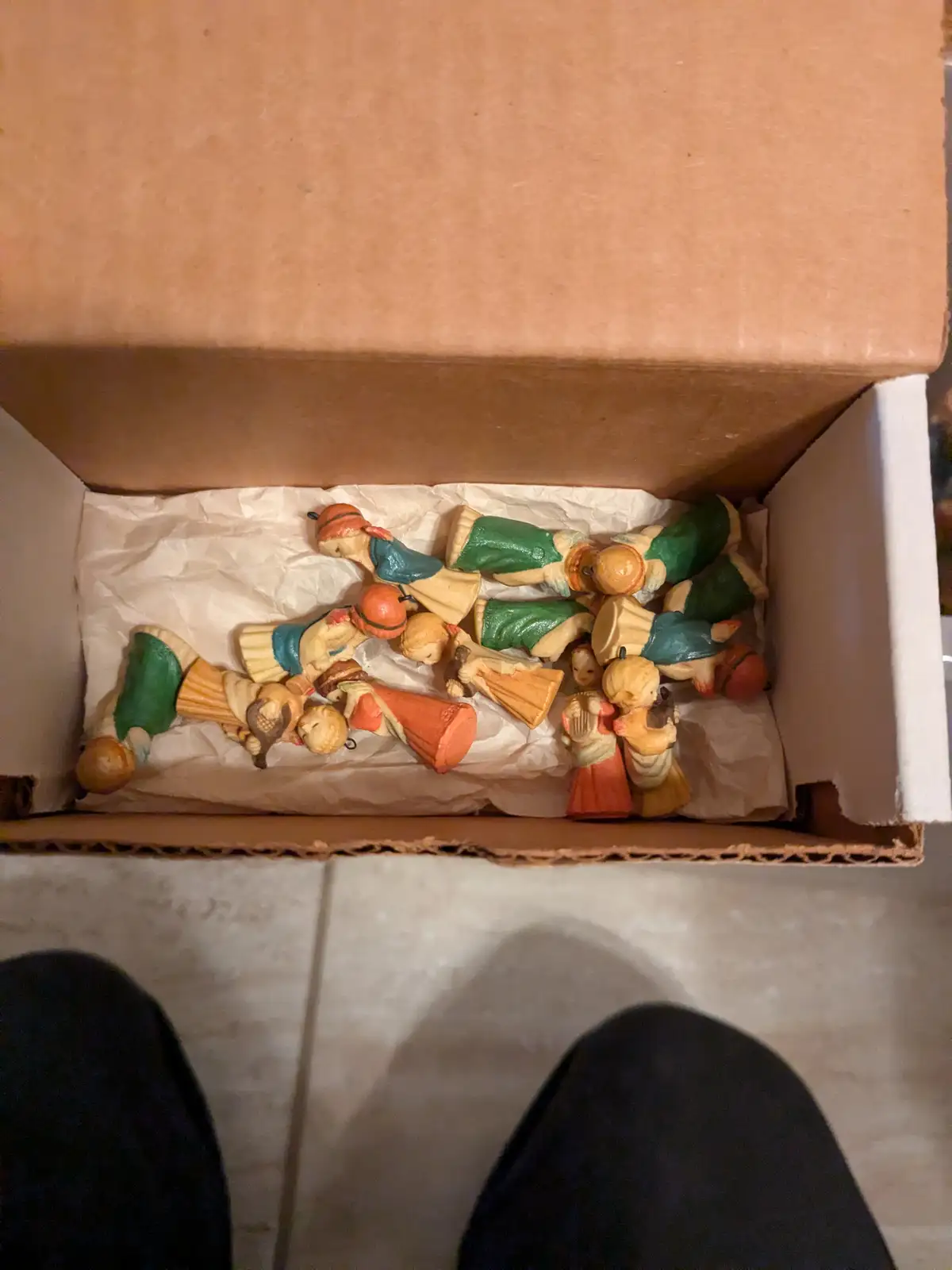 An overhead view shows a cardboard box filled with numerous small, colorful plastic angel figurines, some lying on their…