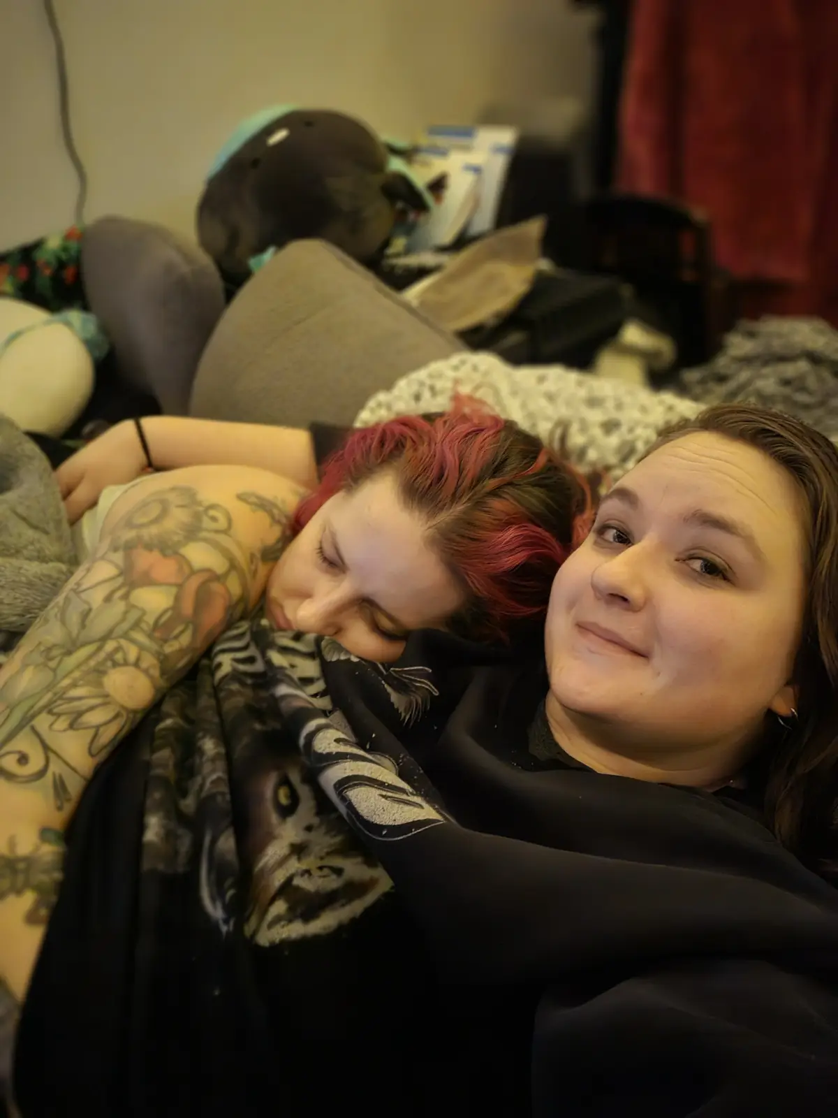 Two people are lying on a couch in a cluttered room; one person with red hair and tattoos is asleep, while the other person…