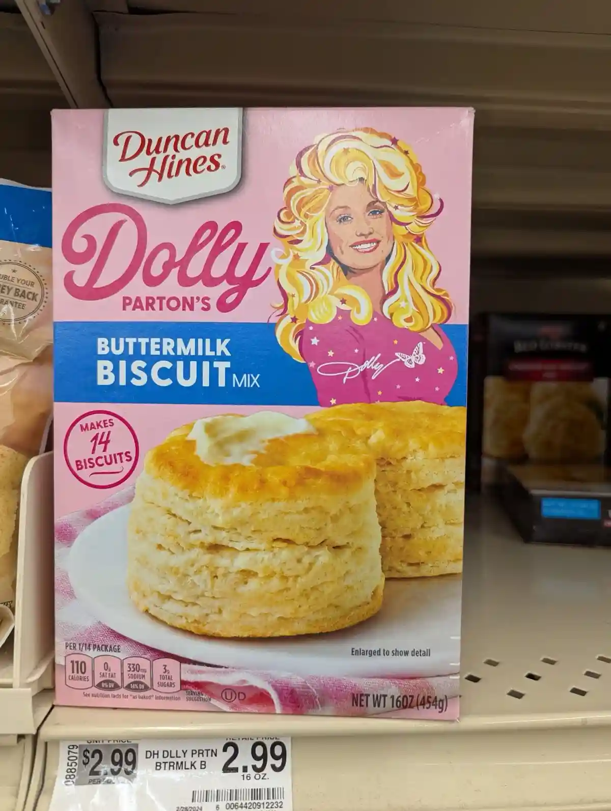 A pink box of Duncan Hines Dolly Parton's Buttermilk Biscuit Mix sits on a grocery store shelf, featuring a cartoon…