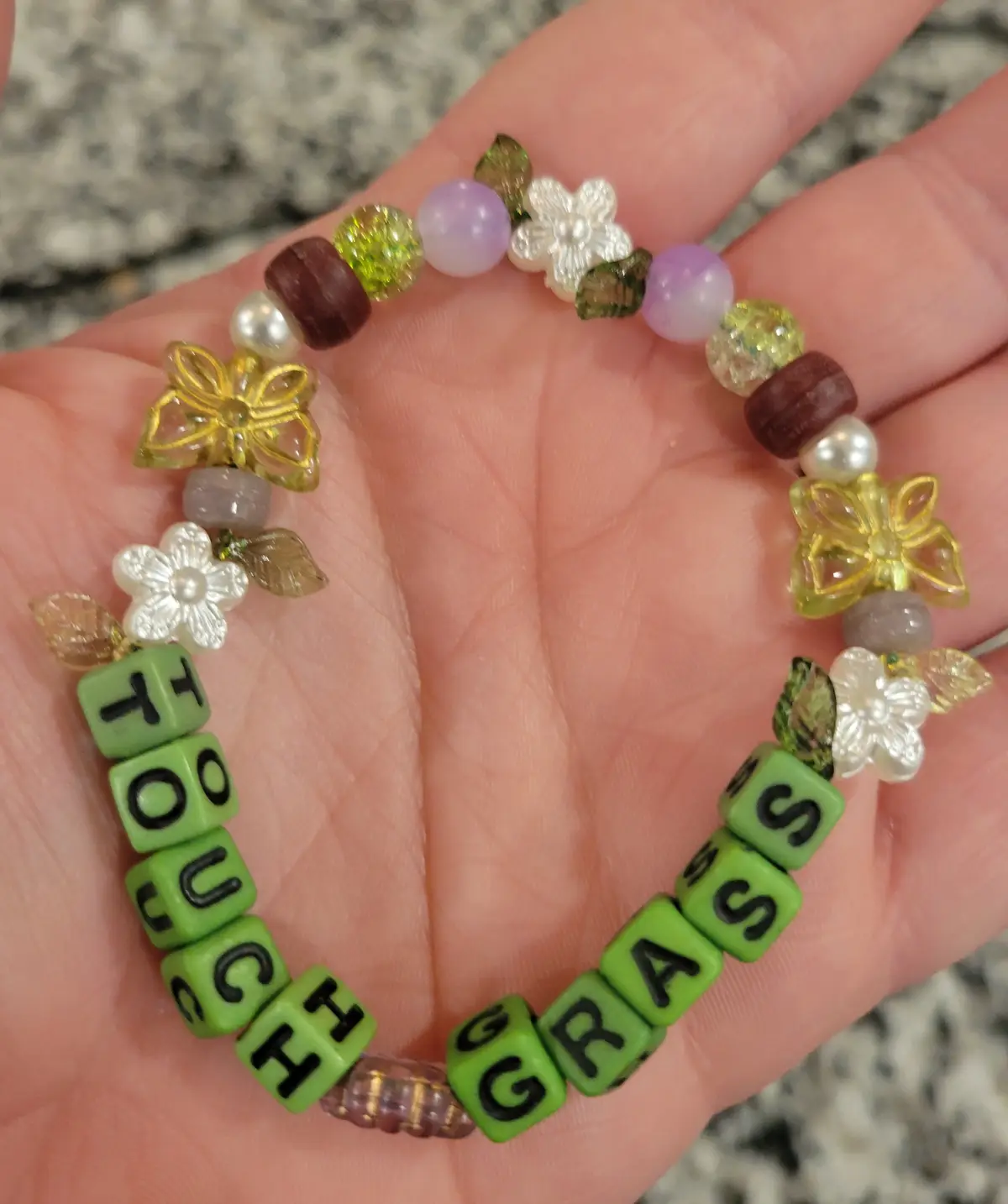 A hand holds a colorful beaded bracelet with green letter beads spelling 'TOUCH GRASS' and decorative flower beads. The…