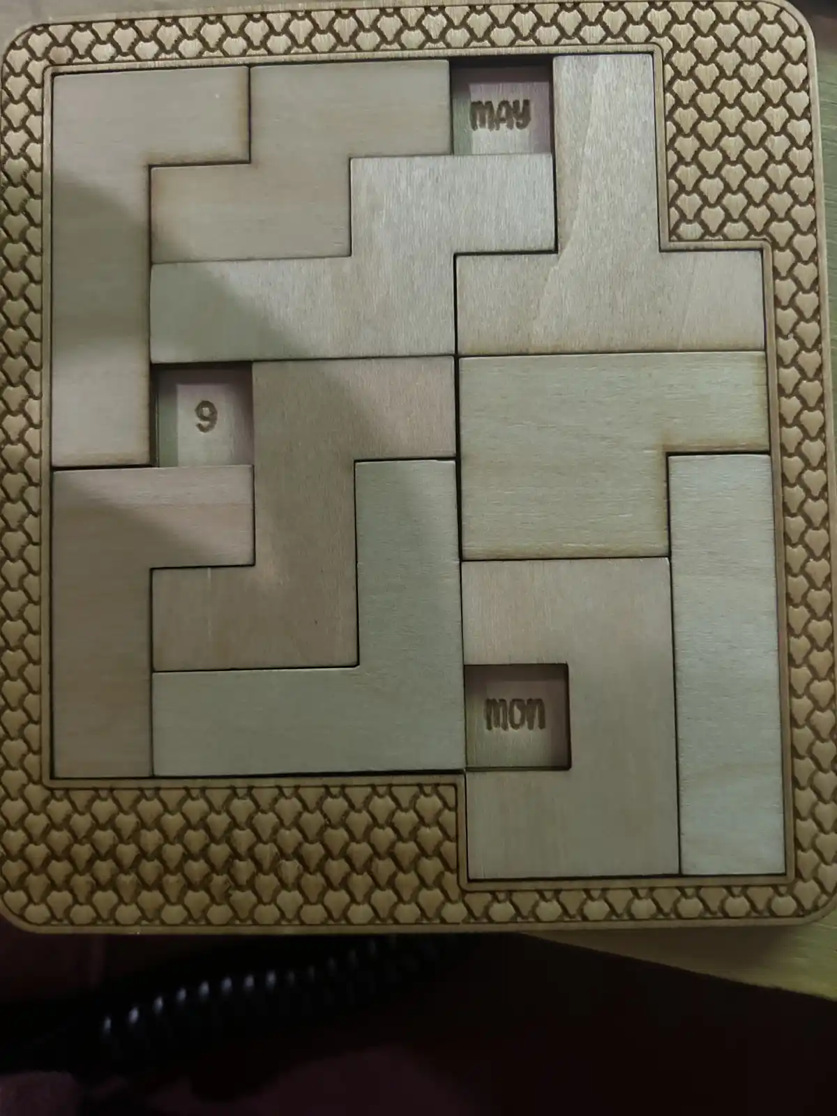 A wooden calendar puzzle with interlocking pieces, showing the month 'may' and the day '9' in recessed slots, with 'mon'…