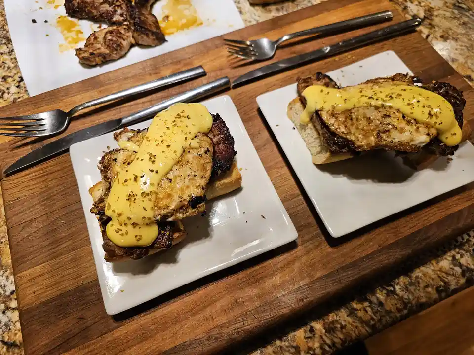 Two plates of food, each featuring a sandwich with a grilled meat patty, a fried egg, and a thick yellow hollandaise sauce…