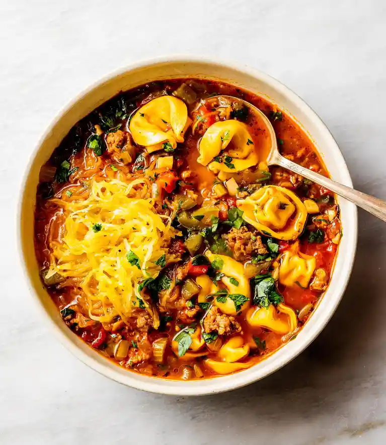 A top-down photo of a white bowl filled with tortellini soup containing meat, vegetables, and parsley, with a large mound of…