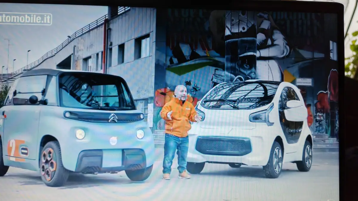 A man in an orange jacket stands between two Citroën Ami electric cars, gesturing enthusiastically. The light blue car on…
