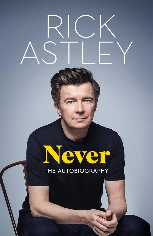 A promotional image of Rick Astley sitting in a chair wearing a black t-shirt with the word 'Never' in large yellow font and…