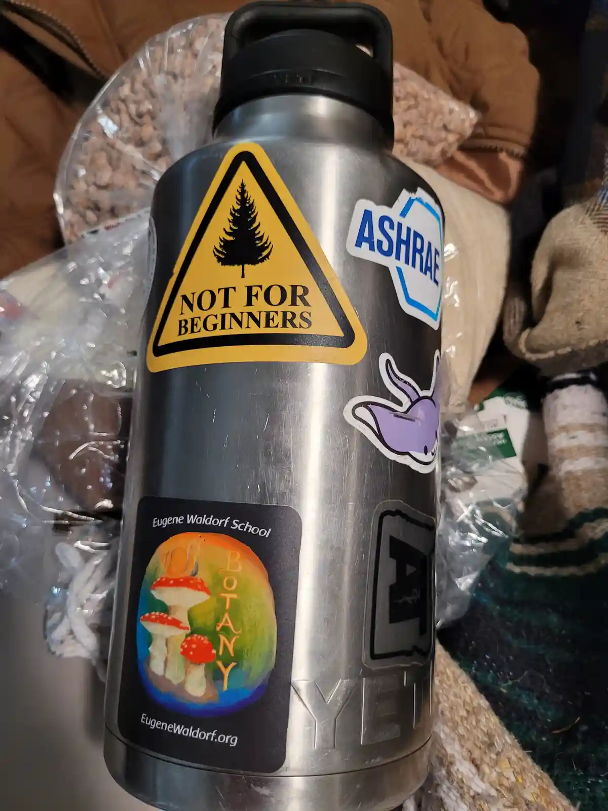 A silver Yeti water bottle covered in stickers, including a prominent yellow triangular warning sign with a pine tree and…