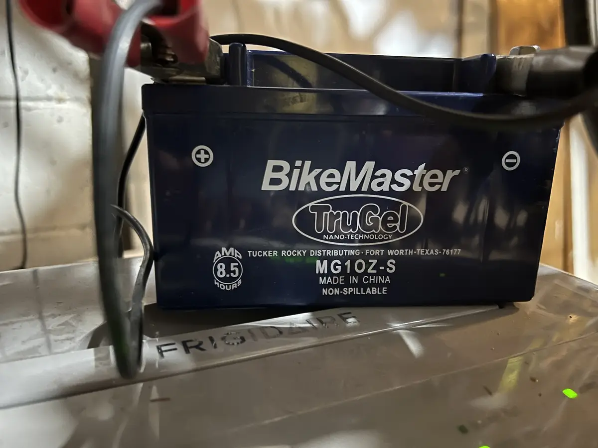 A close-up photo shows a dark blue BikeMaster TruGel motorcycle battery with red and black clamps attached to its terminals…