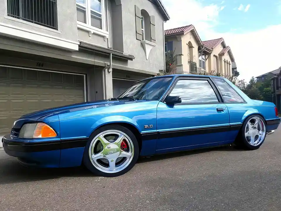 A bright blue 1980s Ford Mustang parked on a residential street in front of a house with a garage door. The car has a…