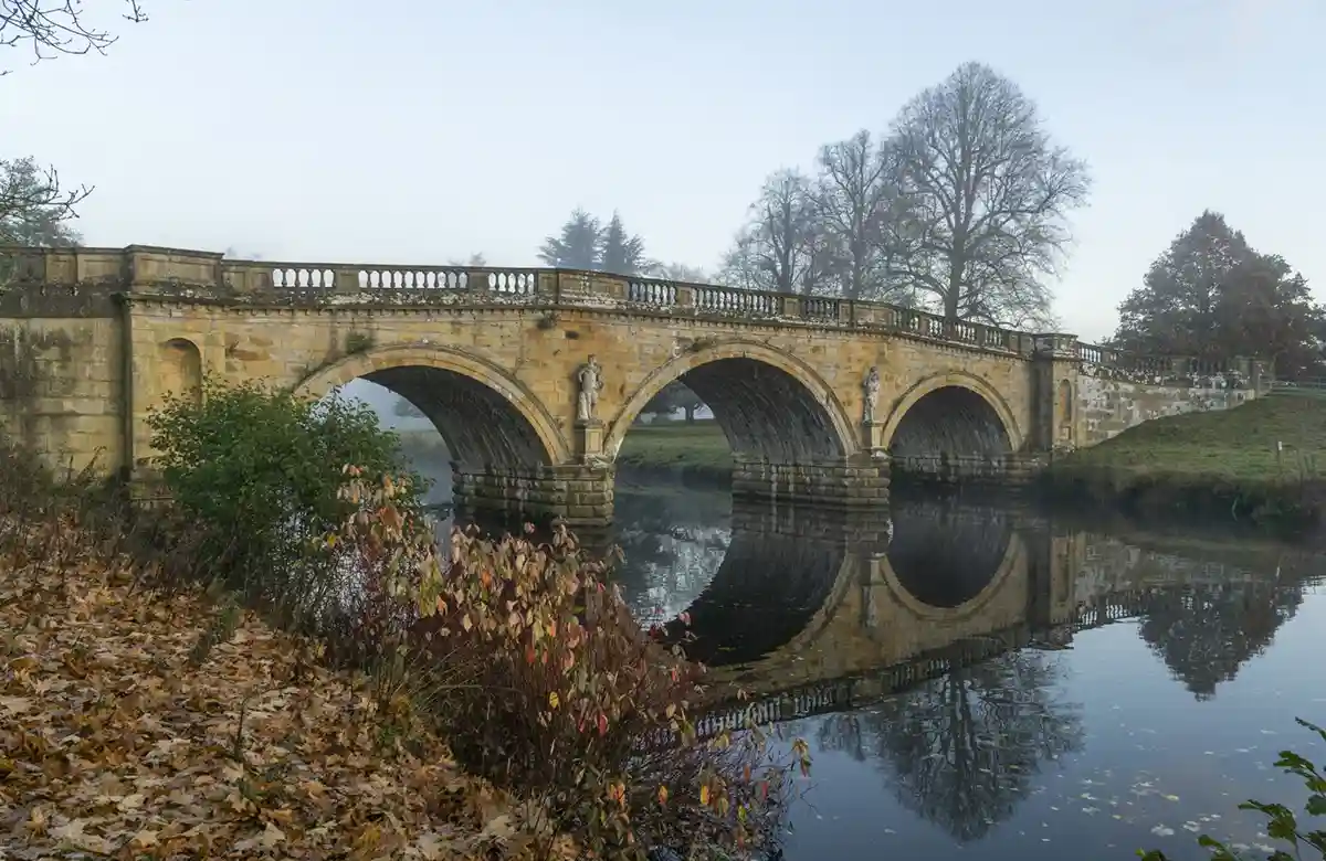 A serene, misty photograph of a stone arch bridge over a calm river, with its reflection perfectly mirrored in the water…