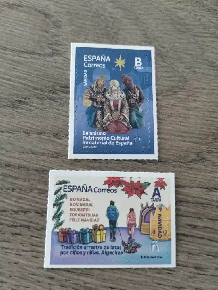 Two Spanish postal stamps from 2024 are displayed on a wooden surface. The top stamp shows a nativity scene with three…
