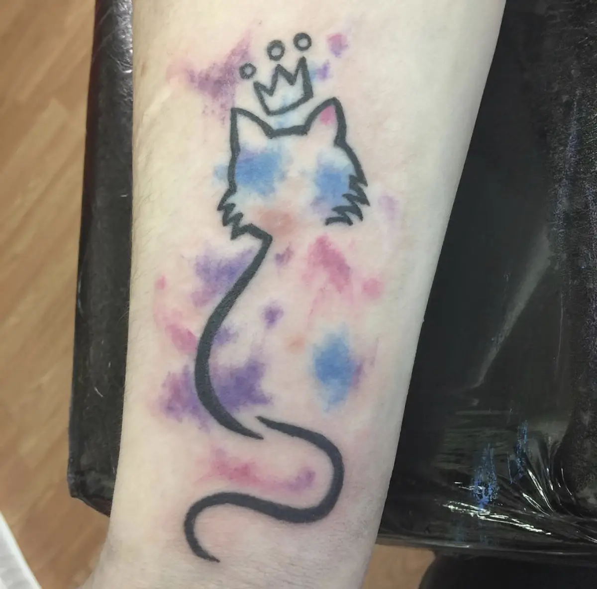 A tattoo on a person's forearm shows a minimalist black outline of a cat's head and long curved tail, with a small crown on…