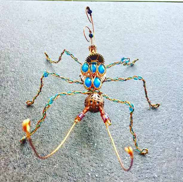 A handmade wire sculpture resembling a spider, crafted from copper-colored wire, blue beads, and a gear, rests on a gray…