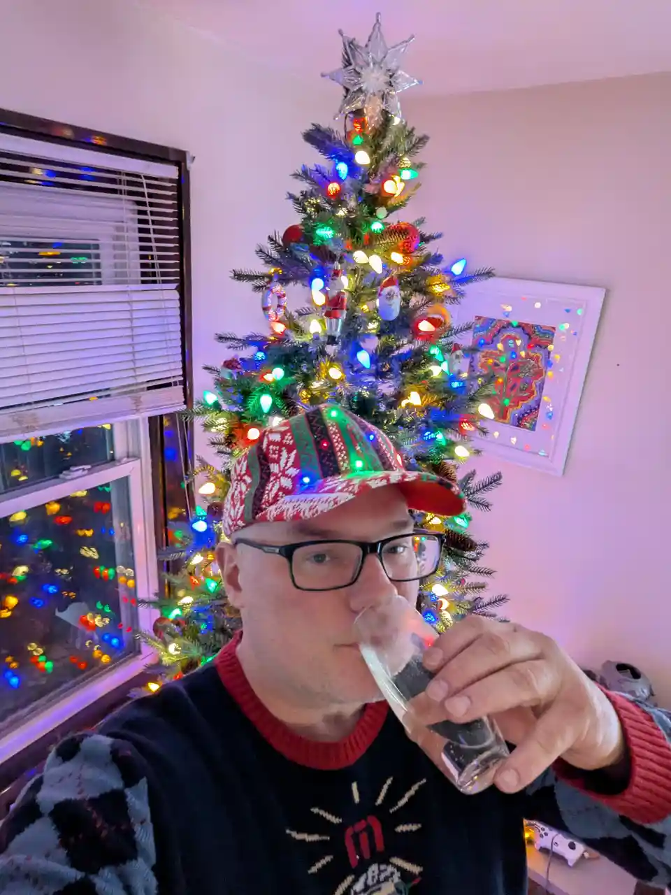 A man wearing glasses, a festive red-and-white patterned hat, and a dark sweater with a red collar drinks from a shot glass…