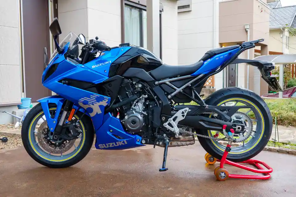 A blue and black Suzuki GSX-8R motorcycle is parked on a wet driveway in front of a house, supported by a red bike stand…