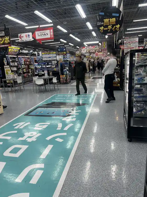 Inside a brightly lit Japanese electronics store, a man walks past a large turquoise floor graphic that reads 'お手洗い'…