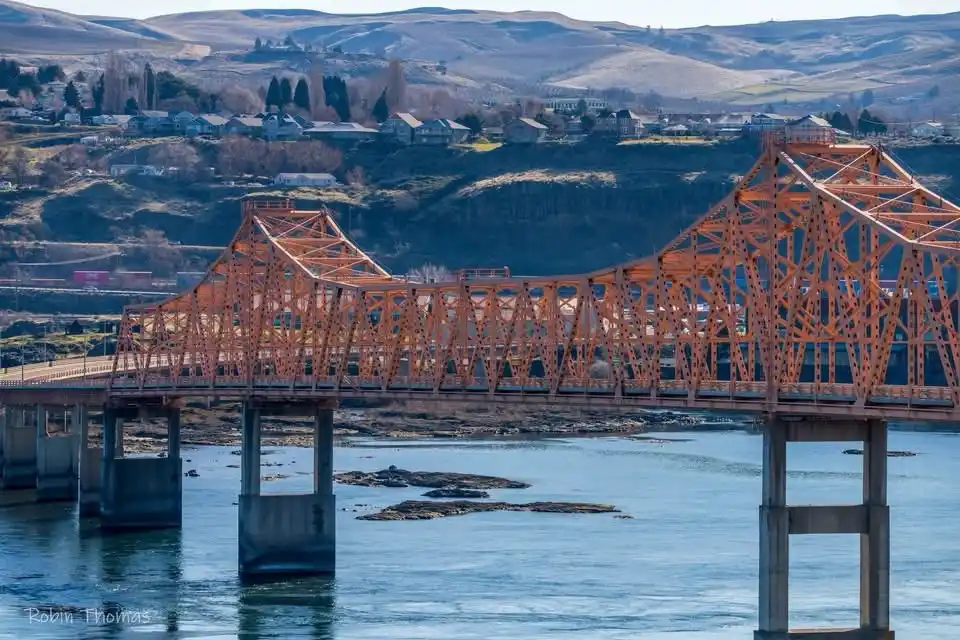 A scenic photograph of a large orange truss bridge spanning a river with rocky outcrops, set against rolling hills and a…