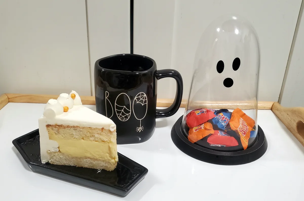 A slice of layered cake with white frosting and gold beads sits on a black plate next to a black mug with 'BOO' and…