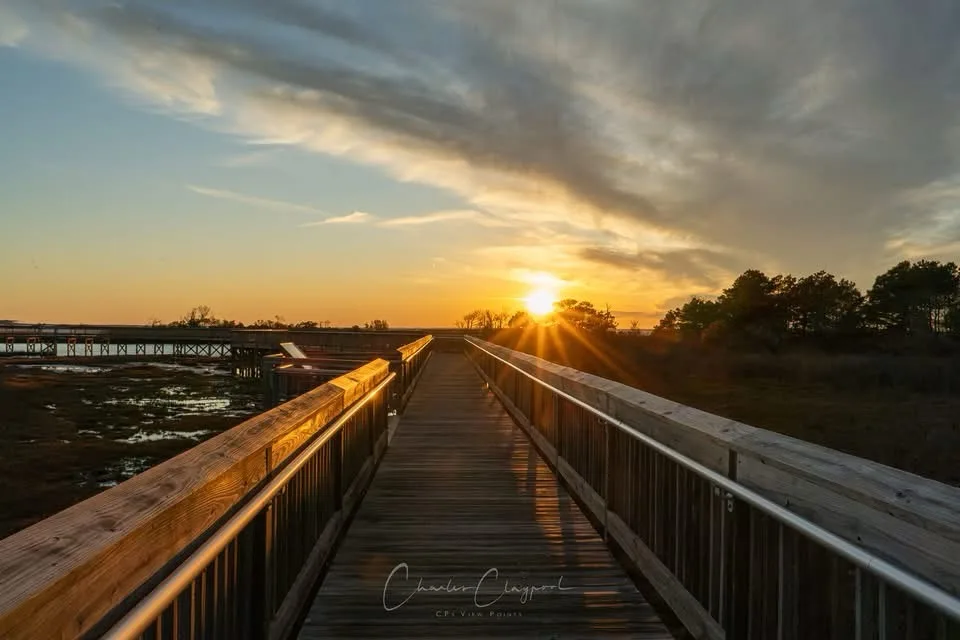 A serene sunset photo shows a wooden boardwalk stretching toward the horizon, with the sun casting golden rays through…