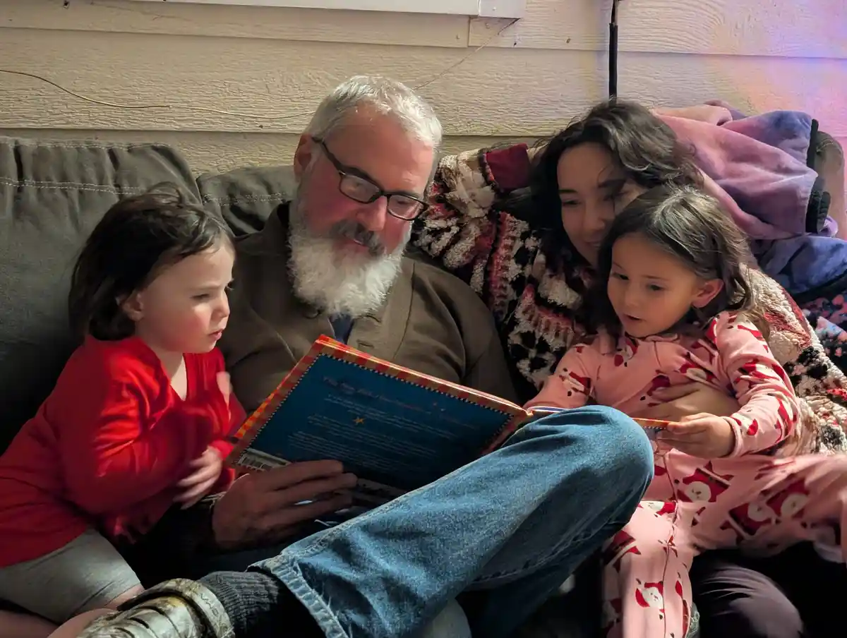A man with a white beard and glasses sits on a couch reading a book to two young girls, one in red and one in pink pajamas…