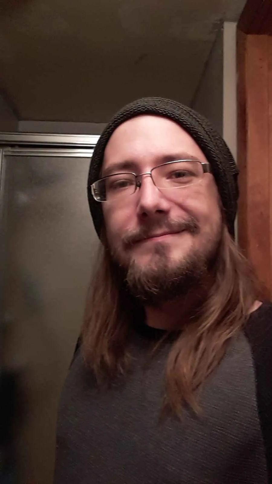 A man with long brown hair, a beard, and glasses, wearing a dark beanie and a dark grey shirt, smiles subtly while looking…