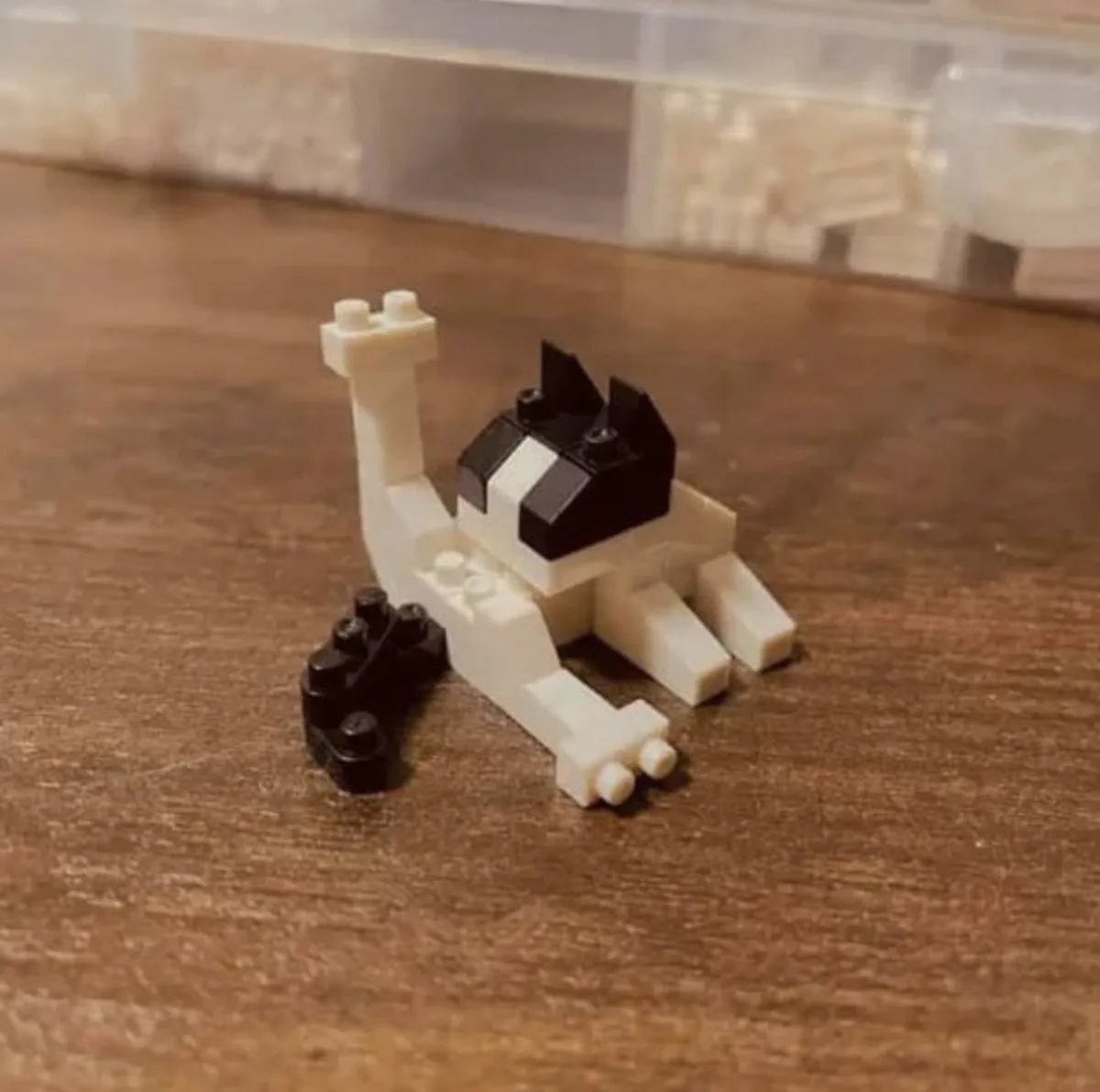 A small, blocky LEGO cat figure with black and white pieces is lying on a wooden surface, appearing to be in a relaxed or…