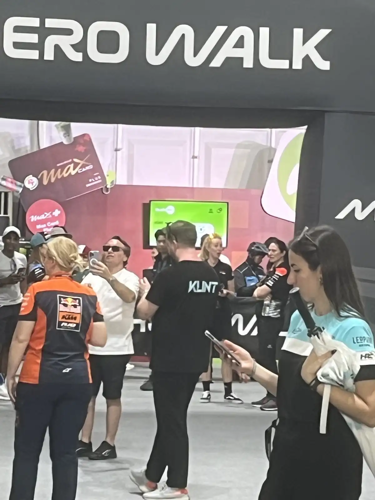 A crowded indoor event space with people milling about, some taking photos, under a large sign that reads 'ERO WALK'. The…