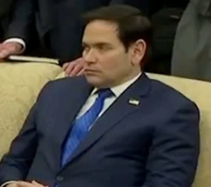 a man in a dark blue suit and blue tie sits on a beige couch with a bored or unimpressed facial expression, looking slightly…