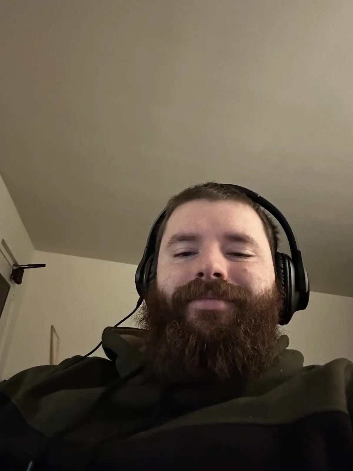 A man with a full beard and black over-ear headphones is looking down with his eyes half-closed, appearing deeply focused or…