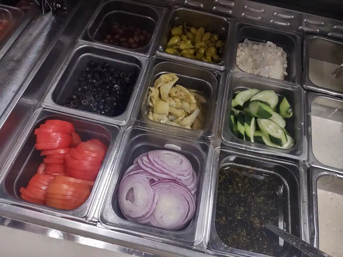 A close-up photo of a salad bar with multiple metal and plastic containers holding various toppings like sliced tomatoes…