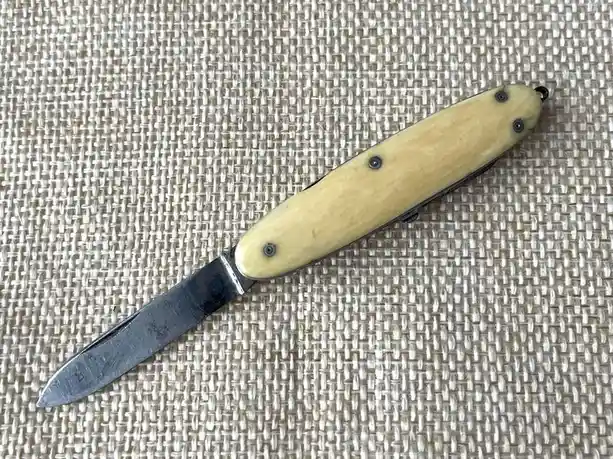 A vintage-style folding pocket knife with a cream-colored, textured handle and a dark metal blade rests on a woven beige…