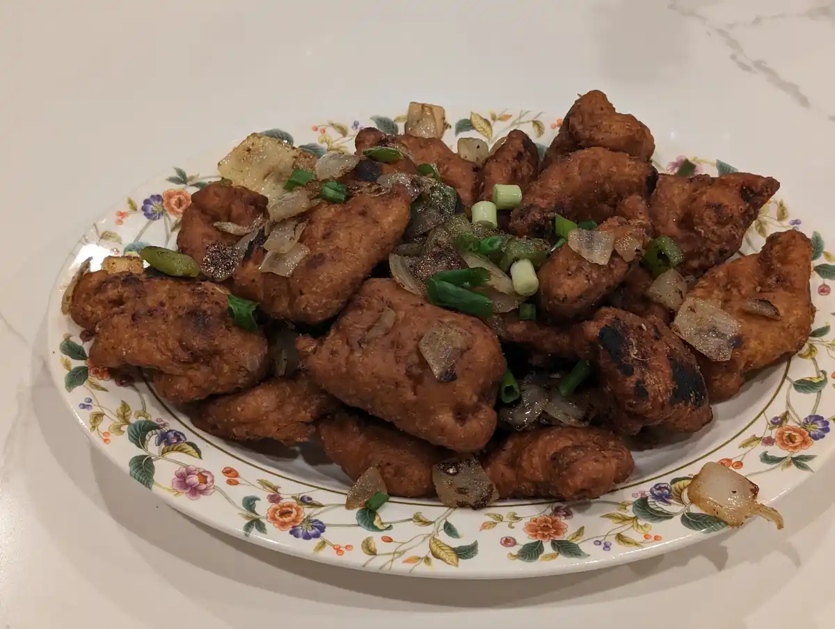 A plate of golden-brown fried chicken pieces mixed with caramelized onions and chopped green onions, served on a…