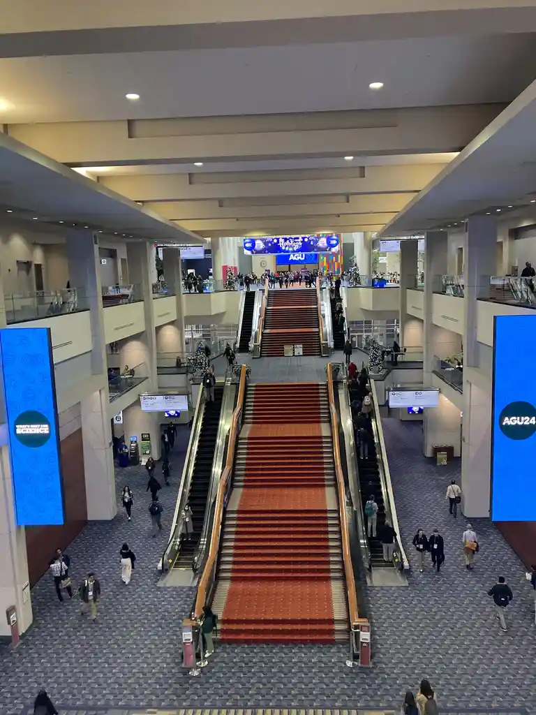 An overhead view of a large, multi-level convention center atrium with escalators and stairs covered in red carpet…