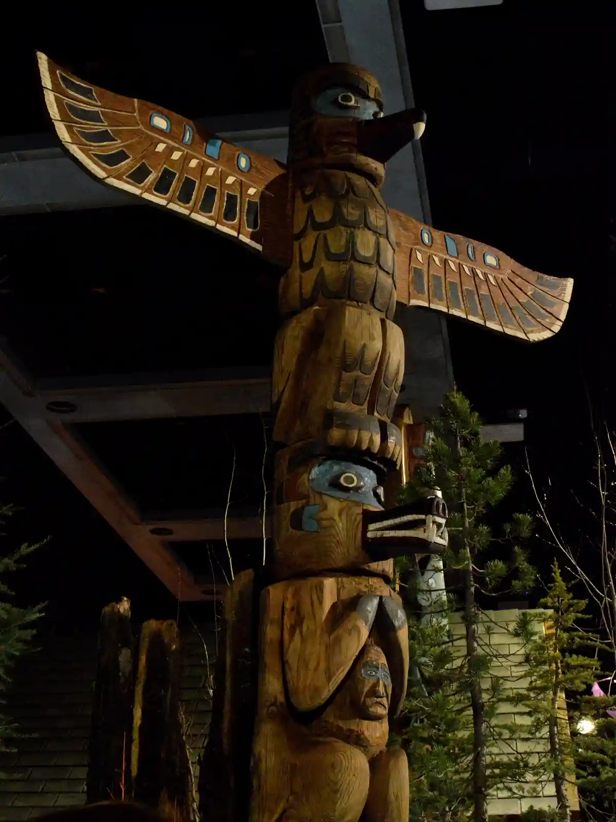 A tall, intricately carved wooden totem pole stands at night, featuring stacked figures including a large bird with…