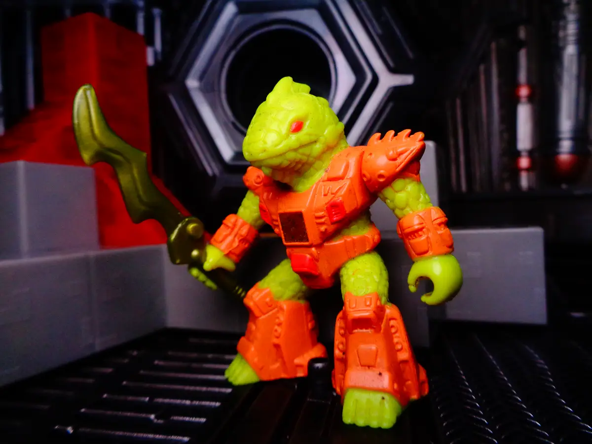 A lime-green, reptilian toy figure with orange armor and a gold weapon stands in a sci-fi setting, overlaid with the text…